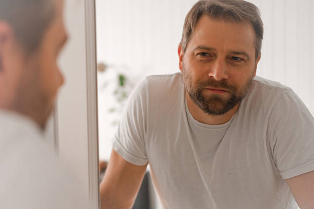 istock Confident handsome bearded man looking himself at the mirror. Pensive guy gazing thoughtfully. Captures introspection, grooming, hair care. Suitable for male health, lifestyle or personal care content 2244950043