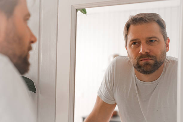istock Confident handsome bearded man looking himself at the mirror. Pensive guy gazing thoughtfully. Captures introspection, grooming, hair care. Suitable for male health, lifestyle or personal care content 2244953426