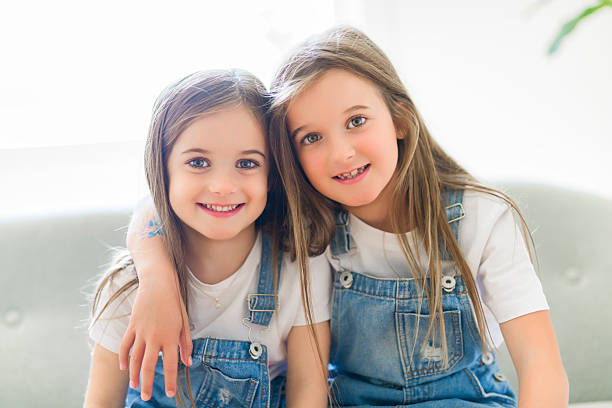 Two sweet little girls stand in their preschool room as they hug and smile for a portrait.  They are both dressed casually and appear happy.