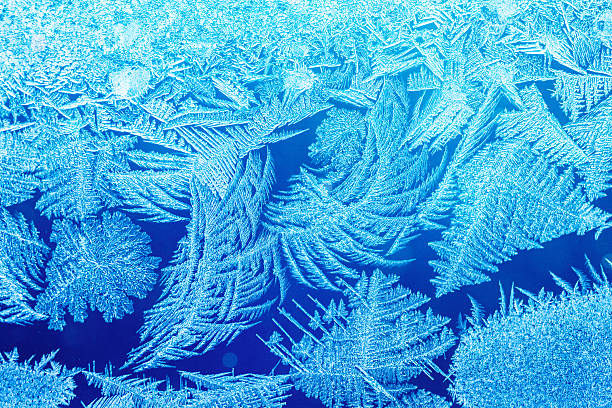 This close-up, high-detail photograph captures the intense, organically grown texture of white and light blue ice crystals and intricate frost patterns covering a transparent surface, likely a pane of glass. The background is a vibrant, deep blue and electric cyan color, with a darker, clear area in the center providing excellent negative space, and the overall effect includes small light particles and a bright glow. This image is excellent for seasonal promotions, winter holiday designs, science topics on freezing, or as a striking, cold-themed abstract background.
