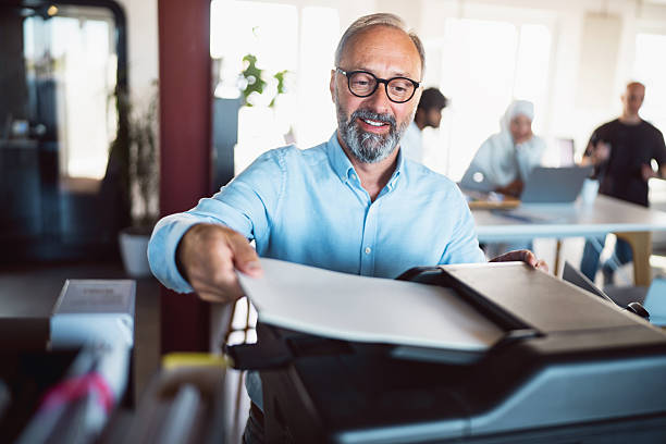 A businessman is seen operating a printer in a contemporary office environment. He appears engaged and focused, contributing to a collaborative workspace where ideas are shared and discussed among colleagues in the background.