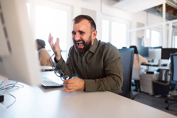 A young business professional working on his computer appears to be angry and shouting, holding his eyeglasses, while people in the background are working on their computers.