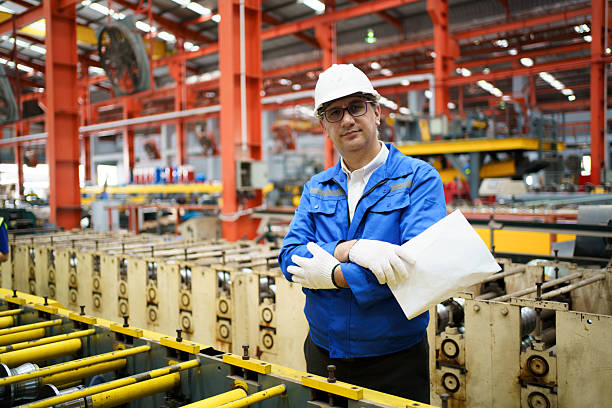 Chief mechanical engineer in work uniform and white safety helmet walking through industrial factory, inspecting machinery readiness and do checklist on documents. Quality control, and industry safety