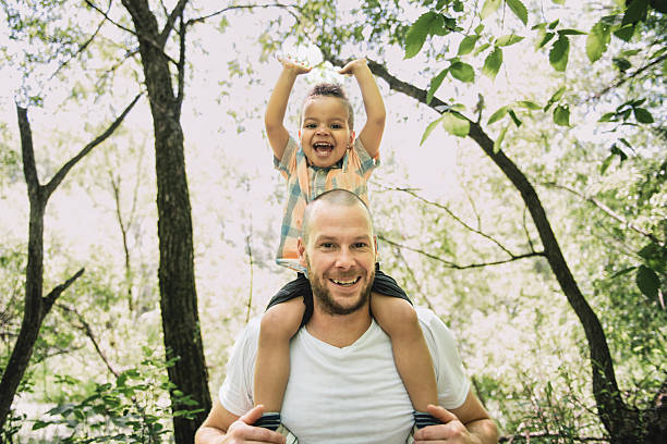A playful father carries his daughter on his back during an outdoor moment, smiling with joy. Warm light and a grassy field backdrop convey active family fun and bonding.