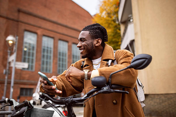 A young blakc man is smiling while using his smartphone, standing next to a bicycle in an urban setting. The backdrop features modern architecture of brick walls typical of Berlin.