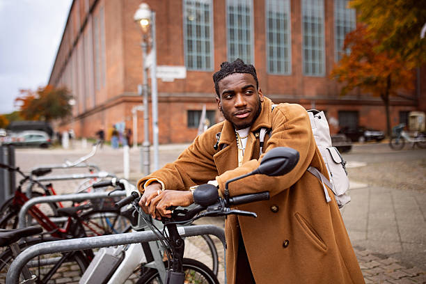 A young man with a backpack leans on a bicycle in an urban setting in Berlin during autumn. The scene captures the essence of city life, showcasing the modern architecture of brick walls and a casual atmosphere.
