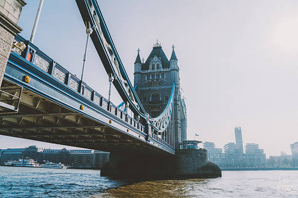 A dynamic upward angle highlights the renowned Tower Bridge with its Gothic towers spanning over the River Thames in London, taken from the bridge’s edge. The iconic Victorian landmark stands against a bright, clear sky with a glimpse of modern city buildings in the distance, capturing a blend of historic charm and vibrant urban life.