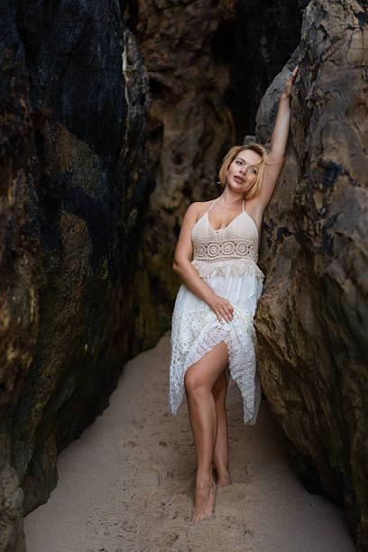 Beautiful blond woman in white lace dress posing between large rocks on sandy beach. Attractive young lady looking thoughtful while leaning on rock