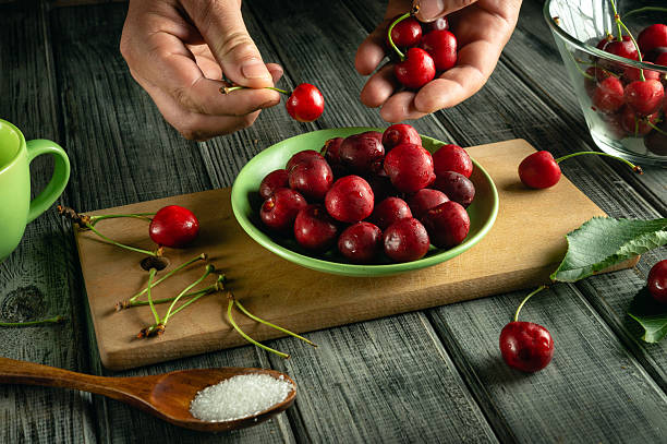 istock Hands carefully remove stems from fresh cherries placed in a green bowl on a wooden board. A cup and sugar sit nearby in a warm, inviting kitchen atmosphere 2244979501