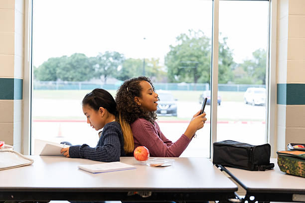 Two young girls sit back-to-back at a table. One girl reads a book, the other uses a smartphone.