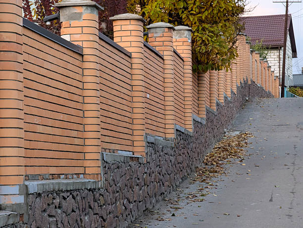 A red brick fence with a stone foundation lines a street that slopes upward, creating a textured urban scene with classic architectural detail.