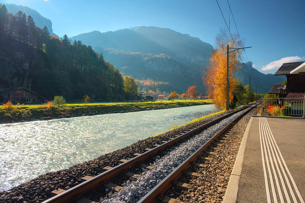 Railway tracks along a river surrounded by autumn trees and mountains in the Swiss Alps. Scenic train route with warm sunlight, small village, and clear blue sky in peaceful alpine valley. Railroad