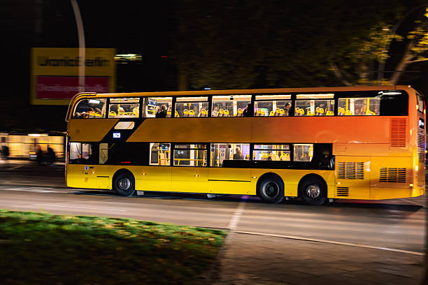 Berlin, Germany – October 15, 2025: Yellow BVG double-decker bus drives through Berlin streets at night, illuminated by street lights. Passengers are visible through the windows.