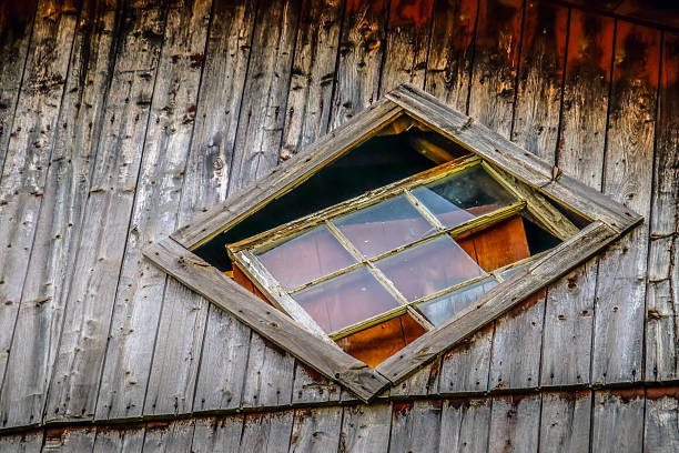 Close-up of a unique diamond-shaped window set into the side of an old wooden barn. The wood is aged and textured with decades of weathering, while the glass panes reflect soft light from the interior. The composition highlights rustic craftsmanship and the timeless charm of rural Quebec architecture.