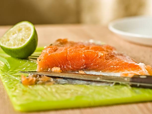 A sharp knife rests on a green cutting board next to a fillet of salted salmon and lime fruit at background