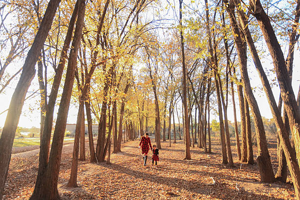Mother and young daughter, wearing buffalo check dresses, walk along pathway in forest, autumn leaves on trees, Rio Grande Valley State Park, Albuquerque, New Mexico