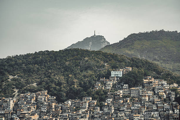 Dense hillside neighborhood in Rio de Janeiro with layered favela houses climbing the slopes, lush green vegetation and distant mountain under hazy daylight
