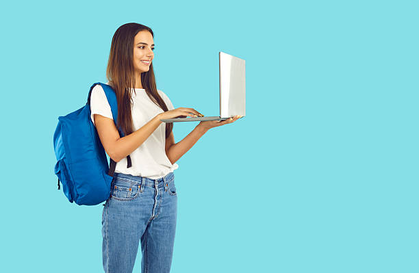 istock Happy student girl with backpack standing on blue background and working on laptop PC 2245029701