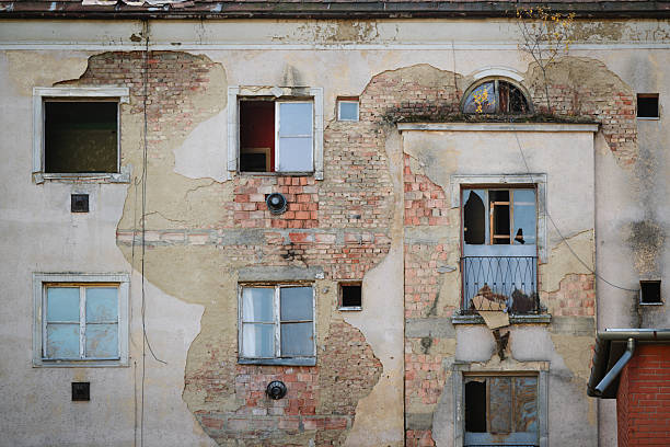 A severely weathered apartment facade shows extensive damage, with crumbling plaster revealing large sections of red and gray brick. Broken windows, missing balcony parts, and faded walls reflect years of neglect and abandonment. The visual texture captures a haunting yet captivating sense of urban decay.