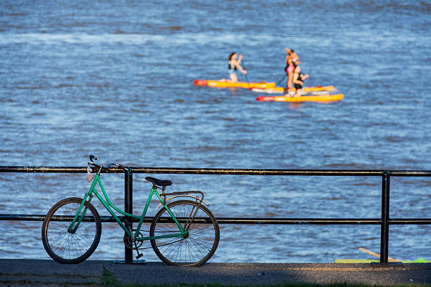 A Vintage bicycle supported on a railing, and three women practice stand up paddle, unfocus behind. under the sun. Photo Date: october 18, 2025. In the city of Rosario, Santa Fe province , Argentina.