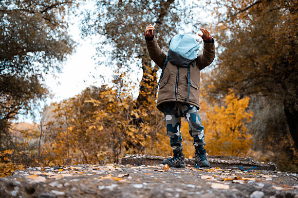 Child hiding face with a light blue hood, playing in an autumn forest filled with colorful leaves on the ground