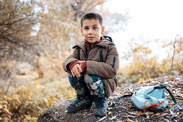 Young boy takes a break during an autumn hike, crouching on a rock with a backpack nearby, experiencing outdoor adventure