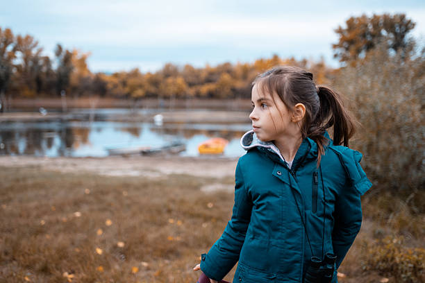 Girl standing by a lake during autumn, looking thoughtfully into the distance. She is wearing a warm jacket, outdoors with binoculars