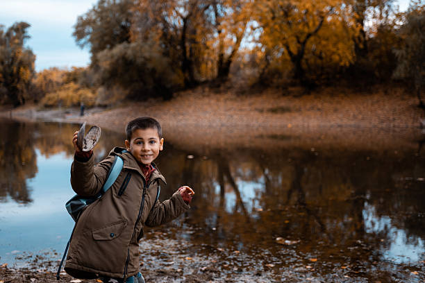 Smiling boy in a brown coat holds a large mussel shell by a calm lake in autumn, exploring nature and enjoying outdoor discovery