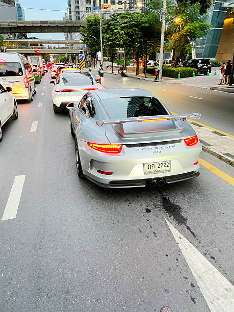 Two types of Porsche cars at stoplight and early evening downtown traffic in Bangkok captured in street Witthayu Road. In foreground is a silver colored Porsche GT3. Ahead is a Porsche Taycan. Cars are standing in queued traffic near Rama I Road. In background are buildings, cars, footbridge. BTS Skytrain  line and a Home Pro sign for a store.