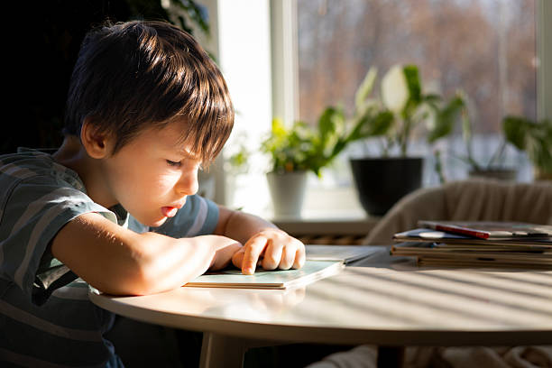 istock Young boy reading a book at a sunlit table, fully focused on learning in a cozy home space 2245066842
