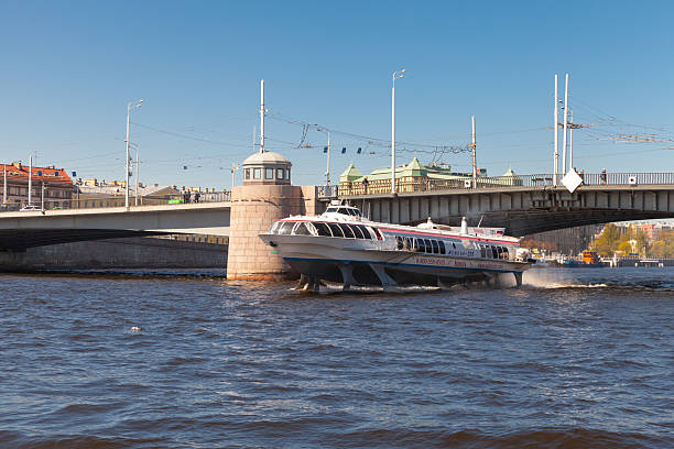 Saint-Petersburg, Russia - May 21, 2022: A sleek speedboat glides under a stone bridge along a broad river, with an urban skyline and green rooftops in the background