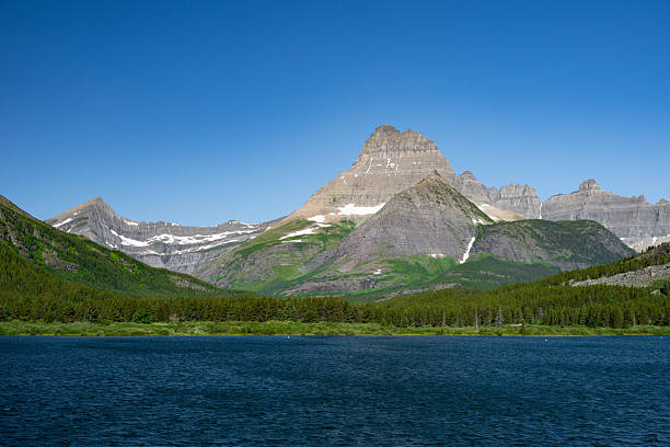 Towering mountain peaks rise above a still alpine lake in Glacier National Park, Montana. The surrounding evergreen forest and vibrant blue sky create a timeless scene of natural tranquility and rugged wilderness.
