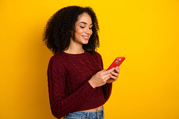 istock Young mixed race woman with curly hair smiles while using red smartphone against bright yellow background showcasing casual fashion shopping vibe 2245090467