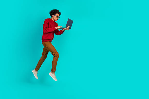 istock Young man in red sweater jumps with laptop against teal background conveying dynamic learning and online shopping vibe 2245090827