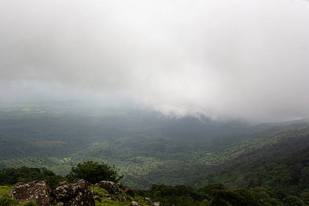 A wide horizontal landscape showcasing the vast, undulating hills and deep valleys of Kerala's high range, heavily draped in mist and fog. The entire scene is covered in dense tropical evergreen forest, creating a monochromatic palette of green and gray. The heavy clouds obscure the peaks and the sky, conveying a sense of seclusion, tranquility, and raw natural beauty on a cold, overcast day.