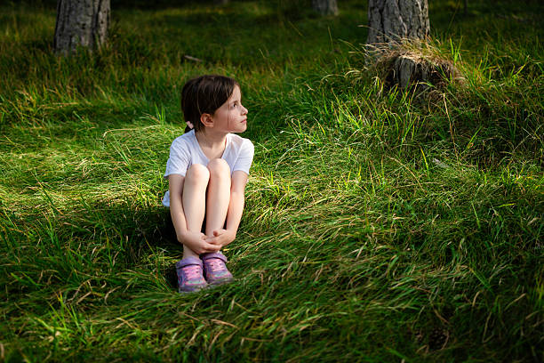 Little girl sitting in green forest grass, looking away thoughtfully, enjoying peaceful summer daydream and quiet childhood exploration