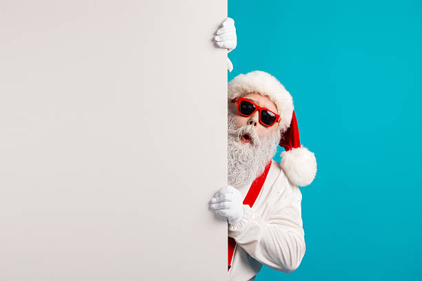 Santa peeks from behind a blank panel with red hat and blue background for a Christmas themed stock photo