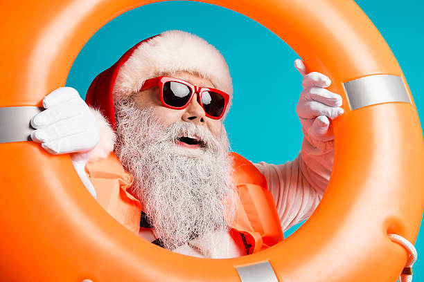 istock Santa with red hat and white beard wears a life jacket and sunglass while holding orange lifebuoy against a vivid blue background 2245098755
