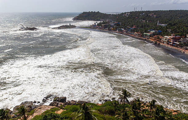 A dramatic, high-angle aerial shot capturing the powerful Arabian Sea as large, foaming waves rush toward the rocky shoreline and a curve of dark sand. The village of Kerala, India, lines the coast, backed by a dense tropical forest of coconut palms. The immense churn of white water in the foreground dominates the scene, conveying the raw power of the ocean on a bright, slightly hazy day.