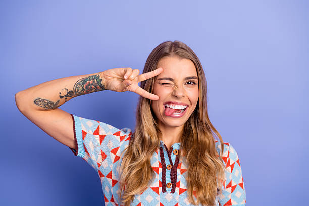istock Young woman with tattoos making a peace sign and playful face wearing a geometric patterned sweater against a blue background 2245099340