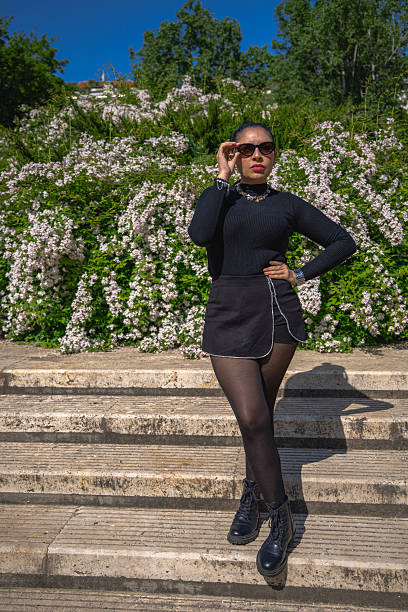 Stylish woman posing confidently on outdoor stone steps. Vibrant blooming bushes and lush greenery create a bright, elegant, and modern fashion portrait under a sunny sky.
