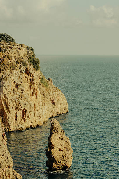 A tall rocky cliff and isolated vertical rock formation rising from the calm blue sea on a clear day, showing natural coastal textures and geological structure.