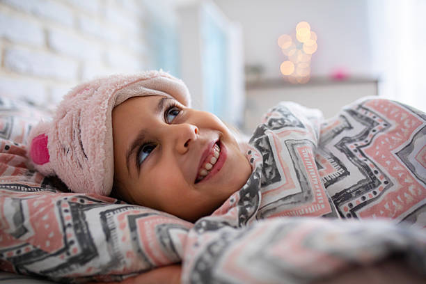 A sleepy little girl lies under her blanket with a pink sleep mask, gently waking up surrounded by soft light and warmth.