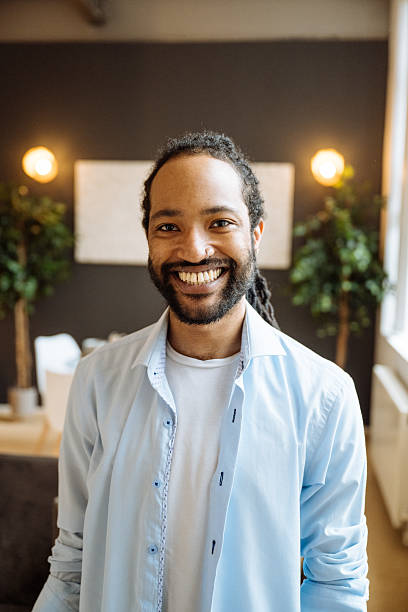 istock Smiling black man with a beard sitting on a dark sofa with surrounded by indoor plants and soft lighting, looking at camera 2245120300
