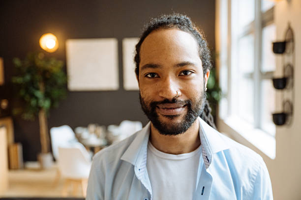 istock Smiling black man with a beard sitting on a dark sofa with surrounded by indoor plants and soft lighting, looking at camera 2245120460