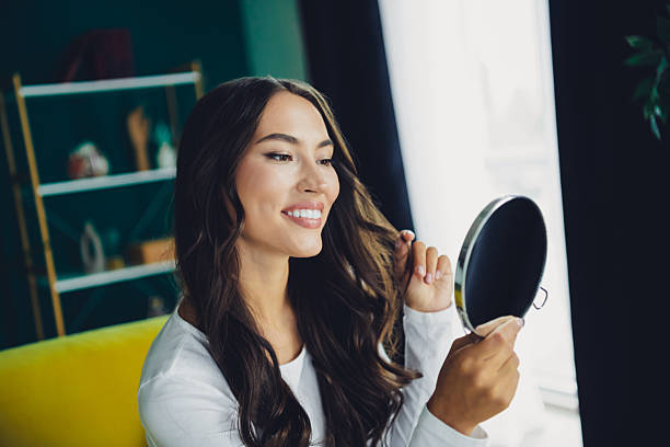 istock Young woman smiles while checking her reflection in a round mirror in a bright home interior with a couch and bookshelf in a cozy living room 2245127771
