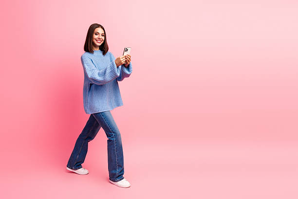 istock Young woman with blue knit sweater walking and smiling while using smartphone against pink studio background 2245128743