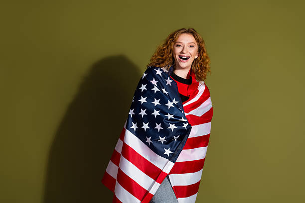 istock Young woman wrapped in American flag smiles brightly in studio setting with olive background selling patriotic fashion lifestyle portrait for advertising and shopping campaigns 2245129696