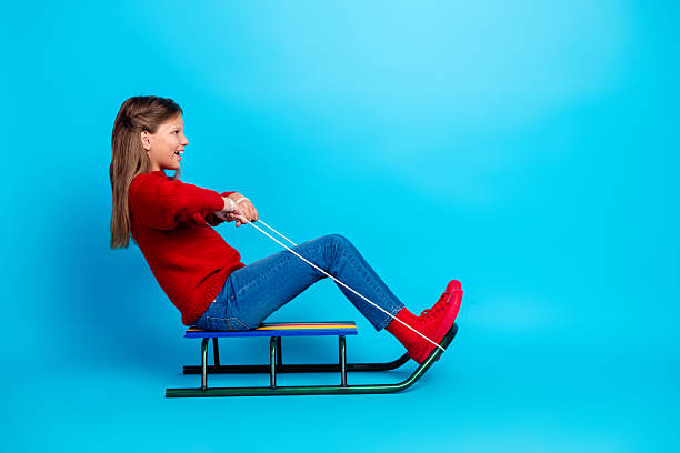 istock Joyful girl on a sled in a bright red sweater with blue jeans on a vivid blue studio backdrop 2245134212