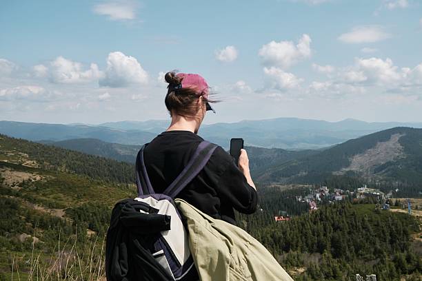 A woman with a pink hat sits on a hill, overlooking the scenic valley in Svydovets mountain range of western Ukraine. She is holding a camera, capturing the picturesque landscape.